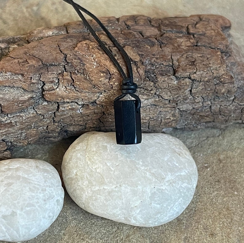 K&ouml;nnte beinhalten: Ein schwarzer Obsidian-Anh&auml;nger an einem schwarzen Lederband. Der Anh&auml;nger ist ein rechteckiger Prisma mit glatten, polierten Facetten.