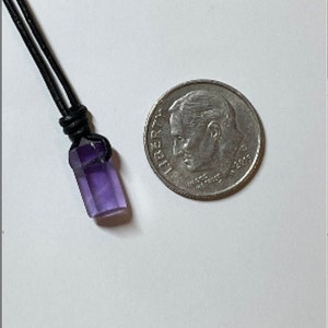 K&ouml;nnte beinhalten: Ein violetter Amethyst-Kristall-Anh&auml;nger an einem schwarzen Lederband. Der Anh&auml;nger ist etwa so gro&szlig; wie eine deutsche Zehn-Cent-M&uuml;nze.