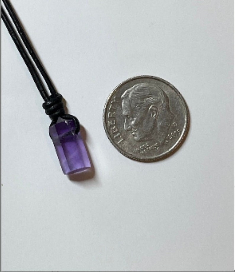 K&ouml;nnte beinhalten: Ein violetter Amethyst-Kristall-Anh&auml;nger an einem schwarzen Lederband. Der Anh&auml;nger ist etwa so gro&szlig; wie eine deutsche Zehn-Cent-M&uuml;nze.