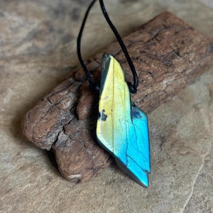 May include: A pendant featuring a polished, irregular shard of labradorite gemstone displaying iridescent yellow and blue flashes, suspended from a simple black cord. The stone has a smooth, faceted surface with visible natural inclusions.