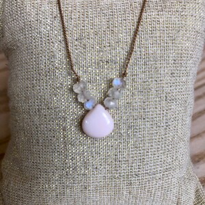May include: A delicate necklace featuring a teardrop-shaped, pale pink pendant. The pendant is surrounded by small, clear, iridescent beads. The necklace has a thin, brown chain. The jewelry is displayed on a textured, light-colored surface.