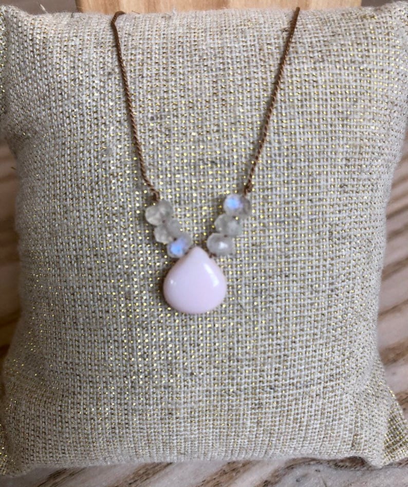 May include: A delicate necklace featuring a teardrop-shaped, pale pink pendant. The pendant is surrounded by small, clear, iridescent beads. The necklace has a thin, brown chain. The jewelry is displayed on a textured, light-colored surface.