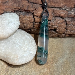 May include: A green moss agate pendant on a brown leather cord. The pendant is a teardrop shape and has a clear, translucent appearance with green moss inclusions.