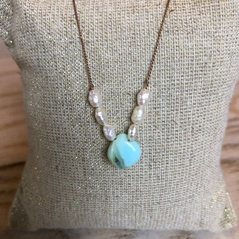 May include: A delicate necklace featuring a light blue, heart-shaped pendant and a strand of white pearls. The pendant is connected to a thin, gold-colored chain. The necklace is displayed on a textured, gold-colored surface.