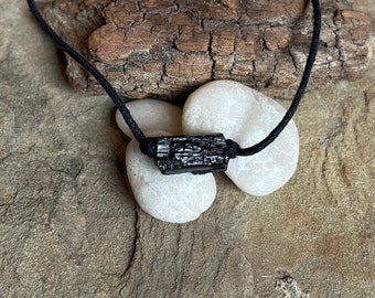 May include: Black Tourmaline crystal pendant on a black cord necklace. The crystal is raw and has a natural, rough texture. The necklace is displayed on a wooden surface with white stones.