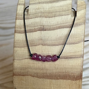 May include: A necklace with five rough, deep pink gemstones threaded on a black cord. The necklace is displayed on a wooden stand, highlighting the natural beauty of the stones.