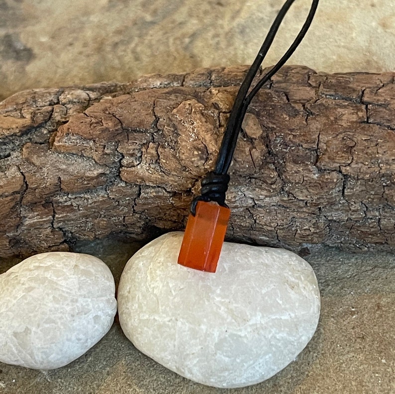 K&ouml;nnte beinhalten: Eine Halskette mit einem kleinen, rechteckigen Anh&auml;nger in Orange auf einem schwarzen Lederband. Der Anh&auml;nger liegt auf einem wei&szlig;en Stein.