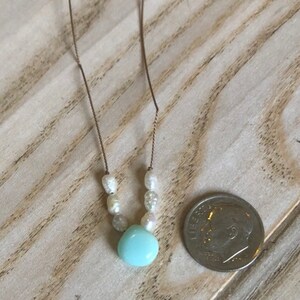 May include: Delicate necklace featuring a light blue pendant and small white pearls. The necklace is strung on a thin, gold-colored chain. A U.S. dime is next to the necklace for size comparison. The jewelry is displayed on a light-colored wooden surface.
