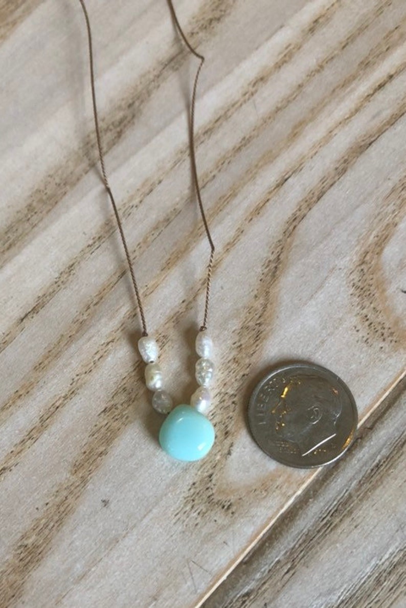 May include: Delicate necklace featuring a light blue pendant and small white pearls. The necklace is strung on a thin, gold-colored chain. A U.S. dime is next to the necklace for size comparison. The jewelry is displayed on a light-colored wooden surface.