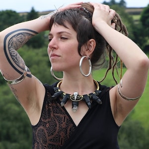 Puede incluir: Un top negro corto con un patrón geométrico marrón y un diseño de envoltura. El top tiene un escote bajo y un detalle de corte en los lados. La modelo lleva un collar plateado con un colgante de cristal y una pulsera plateada en cada muñeca.