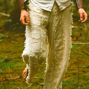 May include: Beige linen pants with a patchwork design and frayed edges. The pants are paired with a cream-colored button-down shirt and brown leather arm cuffs. The image is set in a natural outdoor environment.