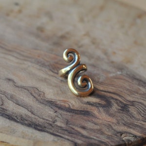 May include: A gold-coloured ear cuff with a swirling, open design. The jewellery piece is set against a wooden background with a natural grain pattern. The cuff appears to be made of metal.