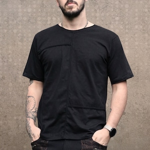Può includere: Un uomo con la barba e gli occhiali indossa una maglietta nera e pantaloni neri. Ha un ciuffo di capelli raccolti in uno chignon e un tatuaggio sul braccio.