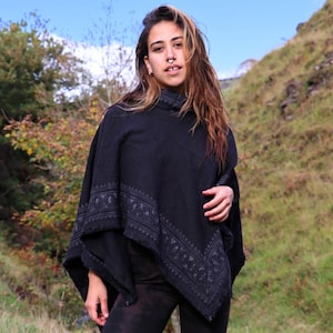 May include: A black poncho featuring a decorative tonal border pattern, paired with black leggings and black lace-up boots. The poncho has a high neckline and a draped silhouette, while the leggings have a subtle mottled texture.