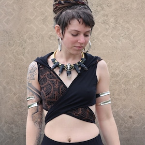 Puede incluir: Top corto negro con diseño envolvente y capucha. El top presenta un diseño estampado marrón en la parte delantera. La modelo lleva brazaletes plateados y un collar con piedras negras y transparentes. El fondo es una pared beige texturizada.