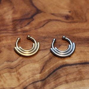 Puede incluir: Dos anillos de nariz, uno dorado y otro plateado, con un diseño en capas. Los anillos están sobre una superficie de madera.