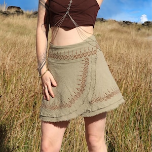 Könnte beinhalten: Eine Frau trägt ein braunes Crop-Top mit einem silbernen Ketten-Body-Harness und einen grünen Wickelrock mit braunen geometrischen Mustern. Sie steht in einem Feld mit hohem Gras vor blauem Himmel.