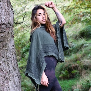 Op de afbeelding: Een olijfgroene poncho met capuchon, een gerafelde zoom en een donker geometrisch patroon langs de randen, gedragen over een donkerpaarse legging. Het kledingstuk heeft een losse, gedrapeerde pasvorm met wijde mouwen.