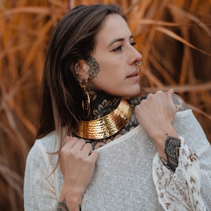 May include: A woman with tattoos and jewellery, wearing a white crocheted top with brown trim. She has a gold choker necklace and large hoop earrings.