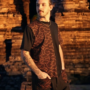 May include: A man with a bun in his hair is wearing a black t-shirt with a brown and orange geometric pattern on the right side. He is standing in front of a stone structure.