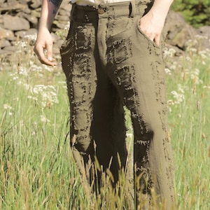 May include: Olive green pants with a distressed, patchwork design. The pants have a classic five-pocket style and a button closure. The model is wearing a cream-colored button-down shirt. The background features a field of tall grass and wildflowers.