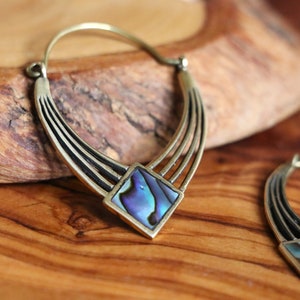 Peut inclure: Une paire de grandes boucles d'oreilles créoles avec un design géométrique. Les créoles sont en laiton et présentent une incrustation carrée en nacre d'ormeau au centre. La nacre d'ormeau a une couleur irisée bleue, verte et violette.