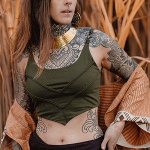 Op de afbeelding: Een vrouw draagt een groene crop top met een gouden choker ketting en een bruin-witte gepatroneerde sjaal. Ze heeft tatoeages op haar armen en borst. Ze staat voor een veld met hoog, droog gras.