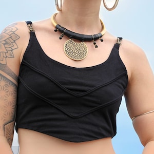Könnte beinhalten: Eine Frau trägt ein schwarzes Tanktop mit einer goldenen und schwarzen Halskette mit einem großen runden Anhänger. Sie hat ein geometrisches Tattoo auf ihrem Oberarm und ein goldenes Armband am Handgelenk.
