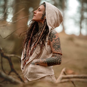 May include: A beige hooded vest with a front pocket. The vest is made of a textured fabric and has a relaxed fit. The person in the image has long, dark hair with reddish-brown dreadlocks and tattoos on their arms and neck.