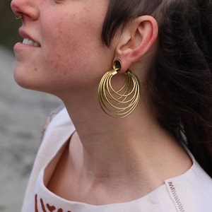 May include: A close-up of a person wearing large, gold-colored hoop earrings. The earrings have multiple nested hoops, creating a layered effect. The person is also wearing a nose ring.