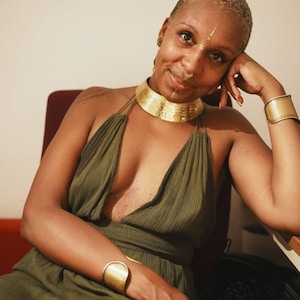 May include: A woman wearing a gold-coloured choker necklace and matching cuff bracelet. She is also wearing a green halter dress. The woman has short blonde hair and a nose ring. She is sitting in a chair.