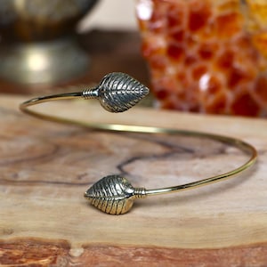 May include: A gold-toned bangle bracelet with two leaf-shaped accents. The bracelet is open and has a simple design.