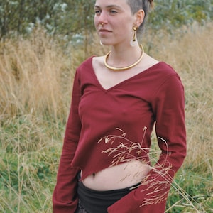Peut inclure: Une femme avec des dreadlocks coiffées en chignon porte une chemise à manches longues rouge bordeaux avec un col en V et un collier ras du cou en or. La chemise est courte et laisse entrevoir son ventre. Elle porte une jupe noire à motifs. La femme est debout dans un champ d'herbe haute.
