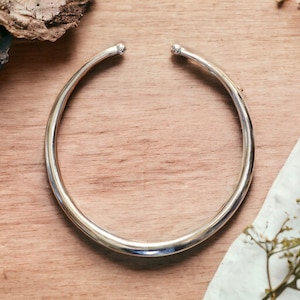 May include: A silver choker necklace with a smooth, rounded surface. The necklace is made of a single piece of metal and has no clasp.