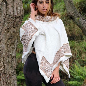 Peut inclure: Un poncho blanc orné d'un motif géométrique marron le long de l'encolure et de l'ourlet. Le vêtement est confectionné dans un tissu texturé à bords effilochés, présentant un motif répétitif de losanges et de chevrons marron sur fond crème.