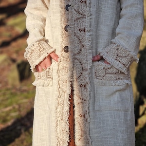 May include: A long, white linen coat with a brown geometric pattern and fringe detailing. The coat has two pockets and a button closure.