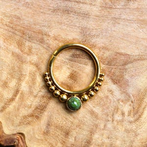 May include: Gold septum ring with a green gemstone and a beaded design.