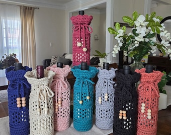 May include: Eight crocheted wine bottle covers in various colours, including blue, beige, pink, teal, grey, black, and coral. Each cover features a crocheted design with wooden beads.