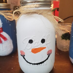 Op de afbeelding: Een wit geverfde glazen pot met een geschilderd sneeuwpopgezicht erop. De sneeuwpop heeft zwarte ogen, een oranje wortelneus en een zwarte glimlach. De pot heeft een bruine touw strik om de bovenkant en een wit papiertje erin.