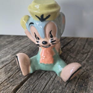 May include: A ceramic figurine of a cartoon mouse wearing a yellow sailor hat, a light blue head, and green overalls with an orange scarf. The mouse has large ears, black whiskers, and a smiling expression, with a glossy finish and visible crazing.