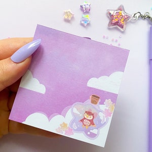 May include: A purple notepad featuring a charming owl illustration inside a bottle. The owl is brown and white with a pink beak. The bottle is surrounded by pink flowers and clouds. The notepad is on a white surface.