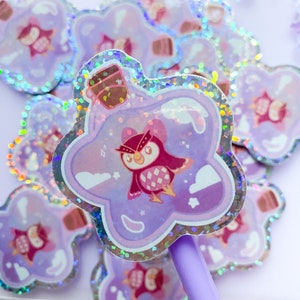 May include: A holographic sticker featuring a purple star-shaped bottle with a pink owl inside. The bottle has a brown cork and is filled with a swirling purple liquid. The owl has a red beak and yellow eyes. The sticker has a glittery finish.