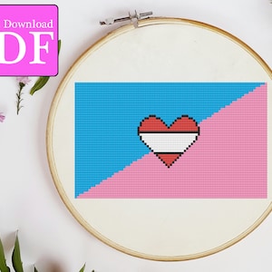Puede incluir: Un patrón de punto de cruz de una bandera del orgullo transgénero con un corazón en el centro. El corazón es rojo con una franja blanca en el medio. La bandera es azul en la parte superior izquierda y rosa en la parte inferior derecha.