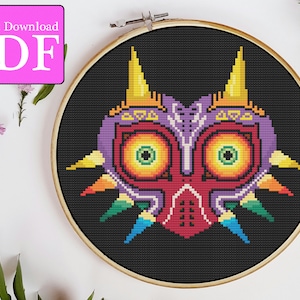 May include: A cross-stitch pattern of Majora's Mask from the Legend of Zelda video game series. The mask is depicted in a pixelated style with a black background and colourful details. The mask has a red, purple, and yellow colour scheme with green eyes.