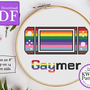 Könnte beinhalten: Kreuzstichmuster für eine regenbogenfarbene Nintendo Switch mit dem Wort "Gaymer" darunter. Das Muster passt auf einen 20 cm großen Rahmen und verwendet 14er Aida-Stoff.