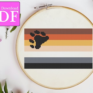 Puede incluir: Un patrón de punto de cruz de la bandera del orgullo de los osos. La bandera tiene seis franjas horizontales en marrón, naranja, amarillo, melocotón, gris y negro. Una huella de pata de oso negra está centrada en las cuatro franjas superiores.