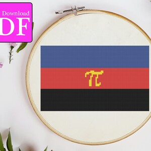 May include: A cross-stitch pattern of a flag with three horizontal stripes: blue, red, and black. The red stripe features a yellow symbol resembling a stylized "T". The pattern is displayed in a wooden embroidery hoop.
