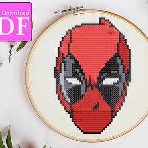 May include: A cross-stitch pattern of Deadpool's face in a red and black color scheme. The pattern is displayed in a circular embroidery hoop.