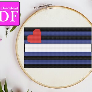 Könnte beinhalten: Ein Kreuzstichmuster einer Pride-Flagge mit einem roten Herzen in der oberen linken Ecke. Die Flagge hat fünf horizontale Streifen: schwarz, blau, weiß, blau, schwarz. Das Herz befindet sich in der oberen linken Ecke des weißen Streifens. Die Flagge befindet sich auf einem hölzernen Stickrahmen.