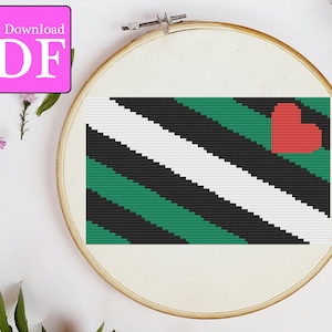 May include: A cross-stitch pattern of a green, black, and white flag with a red heart in the upper right corner. The flag is in a wooden embroidery hoop.